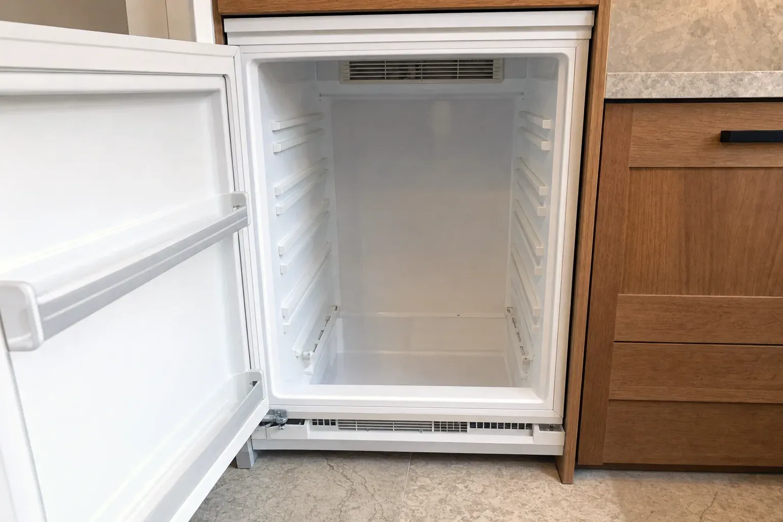 Empty white undercounter refrigerator cavity beside wood cabinet drawers