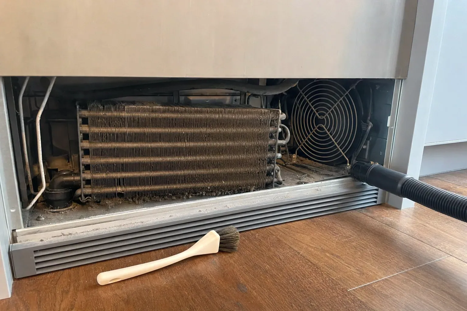 Dust-clogged condenser coil and fan behind a removed refrigerator grille, brush and vacuum hose