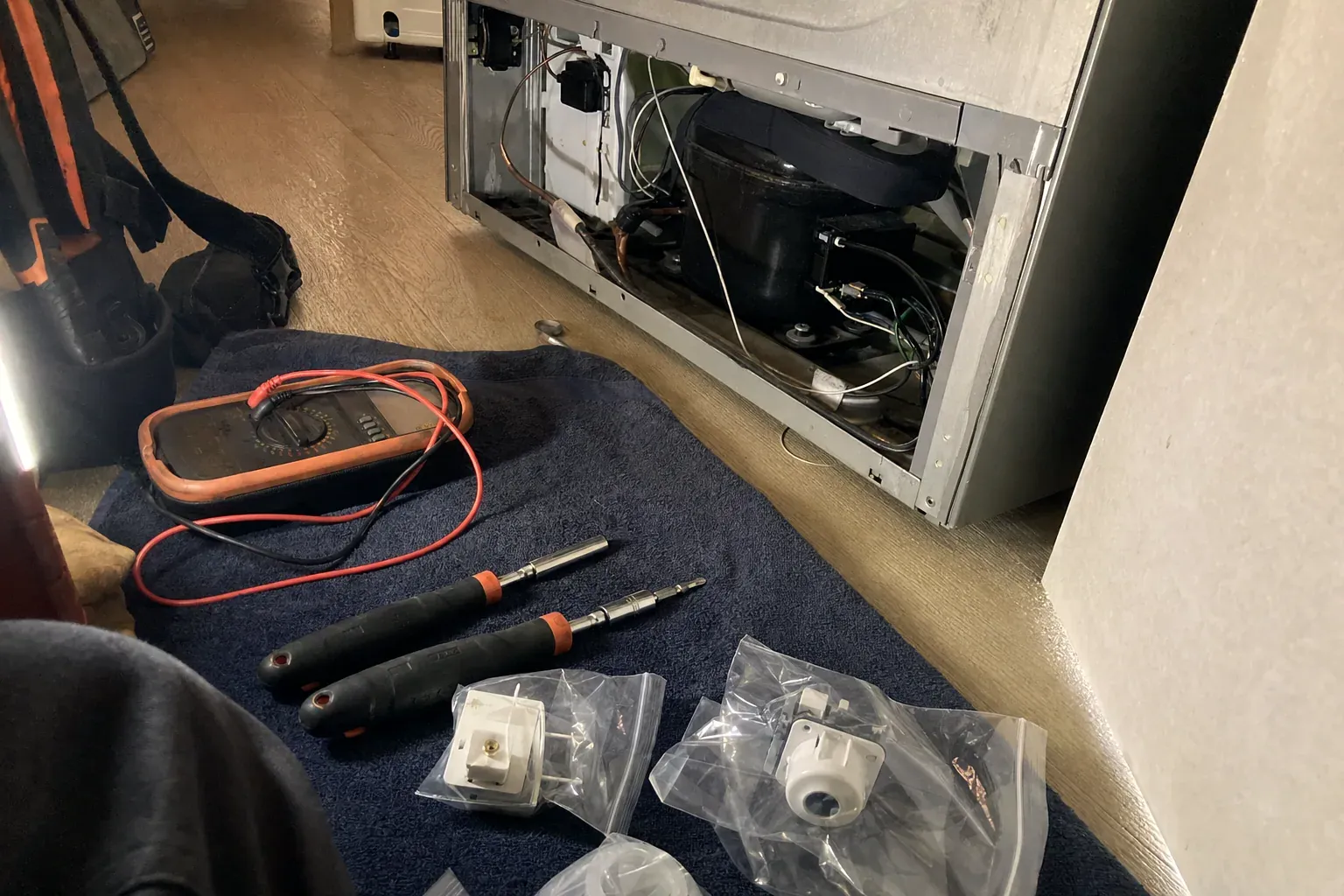 Multimeter, two screwdrivers and bagged replacement parts on a towel beside an open compressor bay