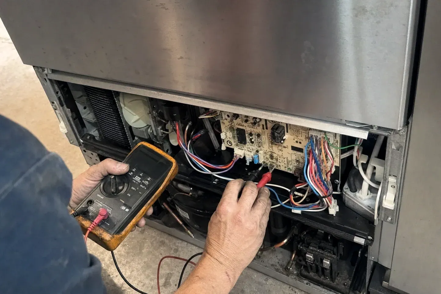 Technician probing a control board with a multimeter behind an open refrigerator panel