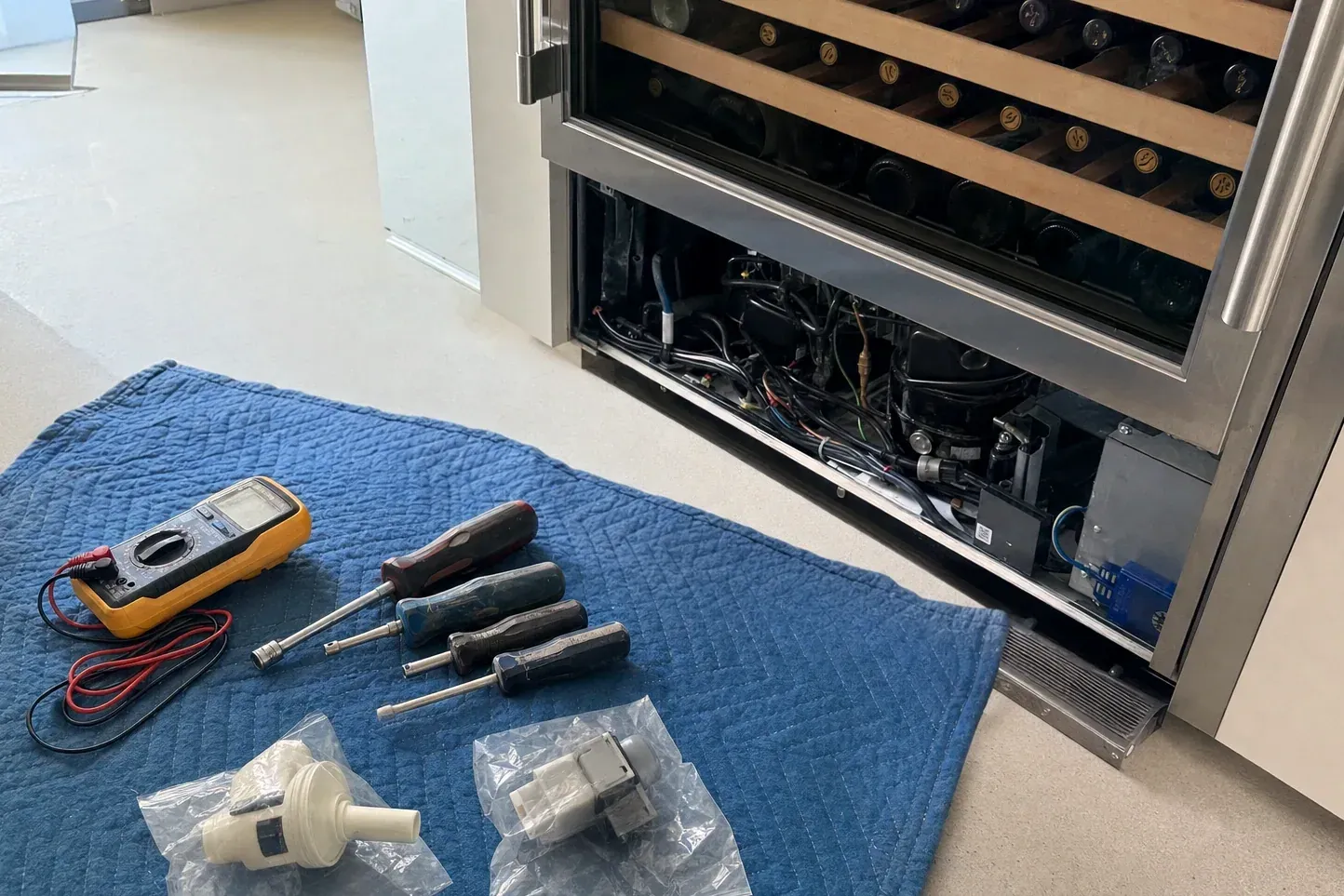 Multimeter, four screwdrivers and bagged valve parts on a blue pad beside an open wine cooler base