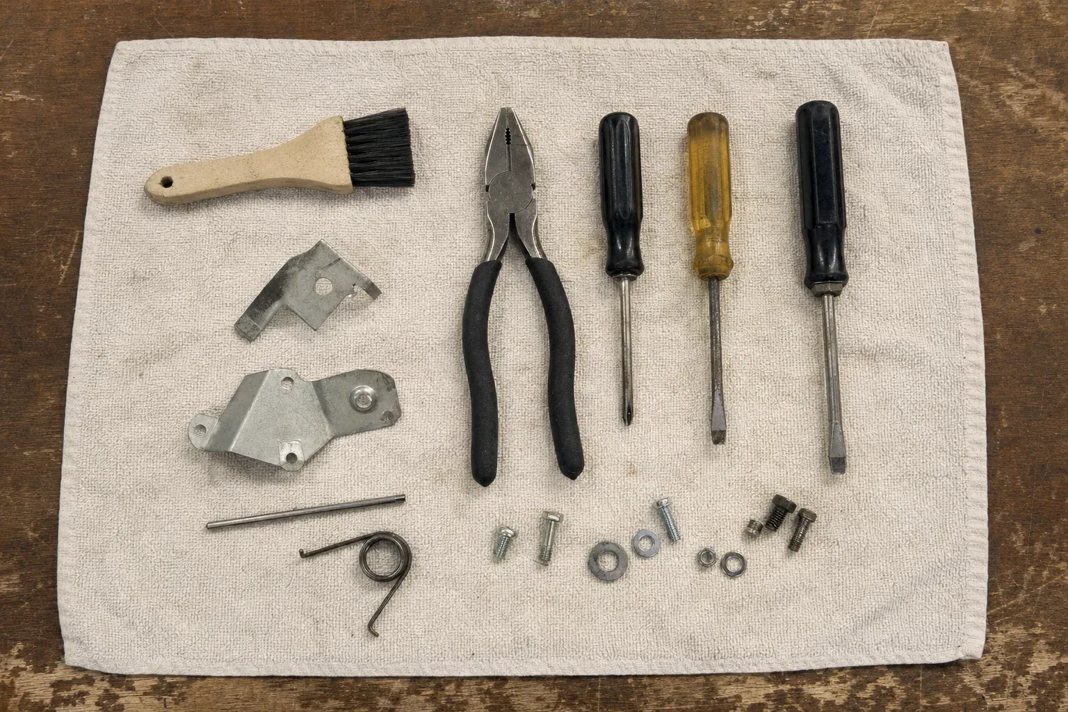 Three screwdrivers, pliers, brush, brackets, spring and hardware on towel